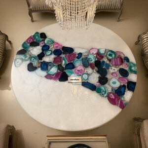 May include: A large, round white table with a decorative inlay of colorful agate stones. The agate stones are in shades of blue, pink, teal, and purple. The table has a glossy finish and is displayed in a room with ornate furniture.