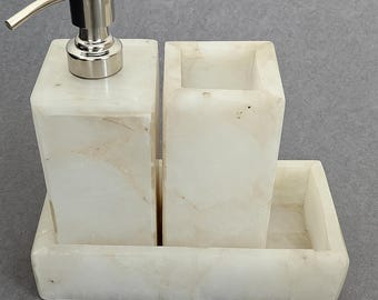 May include: A white marble bathroom set with a soap dispenser, toothbrush holder, and tray. The soap dispenser has a silver pump. The set is made of a light-colored, veined stone, and is designed for bathroom organization.