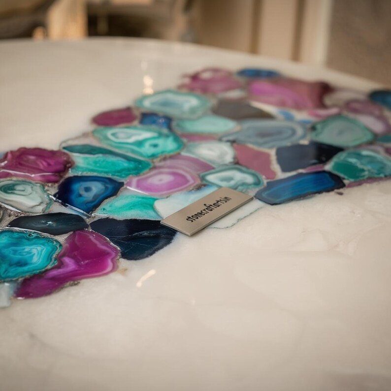 May include: Close-up of a white surface inlaid with colorful agate slices in shades of teal, purple, blue, and gray. A small, rectangular metal tag with the text "stonecraftartsin" is visible. The agate design creates a decorative pattern.
