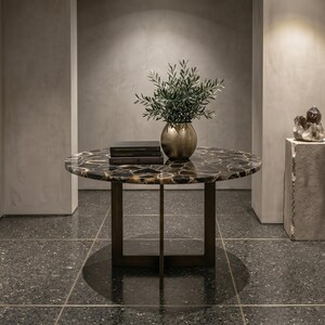 May include: A round table with a dark, polished agate stone top and a bronze-colored metal base. The table features a central cross-shaped support structure. On the surface sits a gold-toned vase containing green foliage and two stacked books.