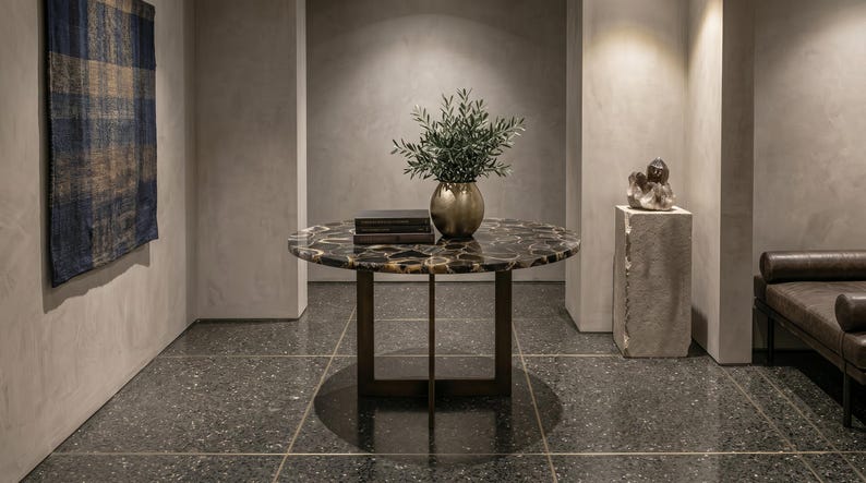 May include: A round table with a dark, polished agate stone top and a bronze-colored metal base. The table features a central cross-shaped support structure. On the surface sits a gold-toned vase containing green foliage and two stacked books.