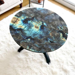 Puede incluir: Mesa de centro redonda con tapa de labradorita. La encimera presenta un patrón arremolinado de azules, verdes y marrones. Se apoya en tres patas negras curvas y descansa sobre una alfombra de pelo largo blanca.