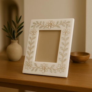 Pode incluir: Uma moldura retangular creme com um design floral. A moldura apresenta uma série de flores e folhas em ouro claro. A moldura está sobre uma superfície de madeira.