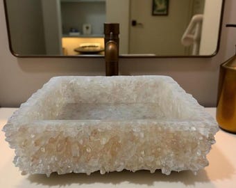 May include: A rectangular, off-white bathroom sink with a textured, crystalline appearance. The sink is set against a neutral background, with a bronze faucet and a matching soap dispenser visible. The sink's unique design adds a touch of elegance.