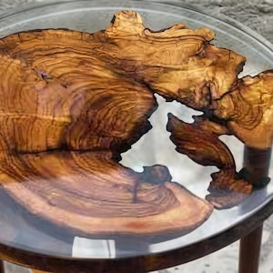 Puede incluir: Una mesa auxiliar redonda que presenta una losa de madera de olivo natural e irregular incrustada en un tablero circular de resina transparente. La madera muestra vetas prominentes y arremolinadas en tonos marrón dorado y ámbar.