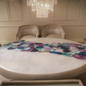 May include: A large, oval coffee table with a white, glossy finish. The tabletop is adorned with a mosaic of colorful, irregularly shaped stones in shades of blue, purple, and teal. The table is surrounded by two armchairs.