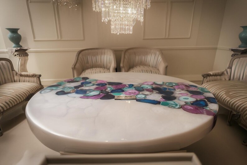 May include: A large, oval coffee table with a white, glossy finish. The tabletop is adorned with a mosaic of colorful, irregularly shaped stones in shades of blue, purple, and teal. The table is surrounded by two armchairs.