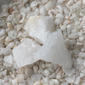 May include: Three large, opaque white quartz crystals rest on a bed of smaller, similarly colored stones. The crystals have a rough, textured surface and varying shapes. The background is filled with numerous small, white pebbles.