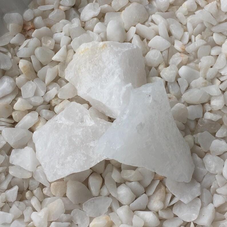 May include: Three large, opaque white quartz crystals rest on a bed of smaller, similarly colored stones. The crystals have a rough, textured surface and varying shapes. The background is filled with numerous small, white pebbles.
