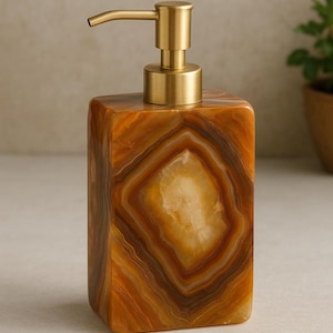 Puede incluir: Un dispensador de jabón rectangular con una bomba dorada. El dispensador está hecho de una piedra marrón y naranja con un patrón en capas. Es un accesorio decorativo para el baño.