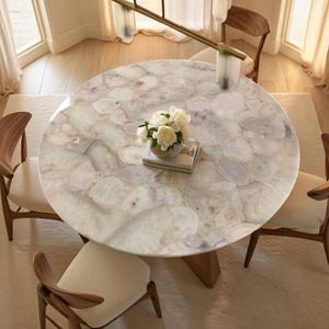 May include: A circular dining table with a white and grey patterned surface, supported by a wooden base. Four wooden chairs with cream-coloured seats surround the table. A vase of white flowers and a book are on the table.