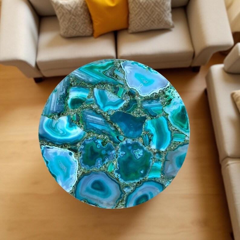 May include: A round coffee table with a mosaic top made of blue and green agate stone. The table's surface is a vibrant mix of colors and patterns, creating a unique and eye-catching design. The table is a decorative piece for a living room.