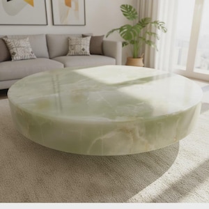 May include: A large, round coffee table with a polished, light green onyx surface. The table is low to the ground and has a solid, cylindrical base. The room has a neutral color palette with a gray sofa and beige carpet.