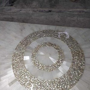 Puede incluir: Una mesa de mosaico con un dise&ntilde;o circular. El centro presenta anillos conc&eacute;ntricos de material brillante e iridiscente. La parte exterior de la mesa est&aacute; hecha de baldosas rectangulares blancas, que irradian hacia afuera desde el centro.