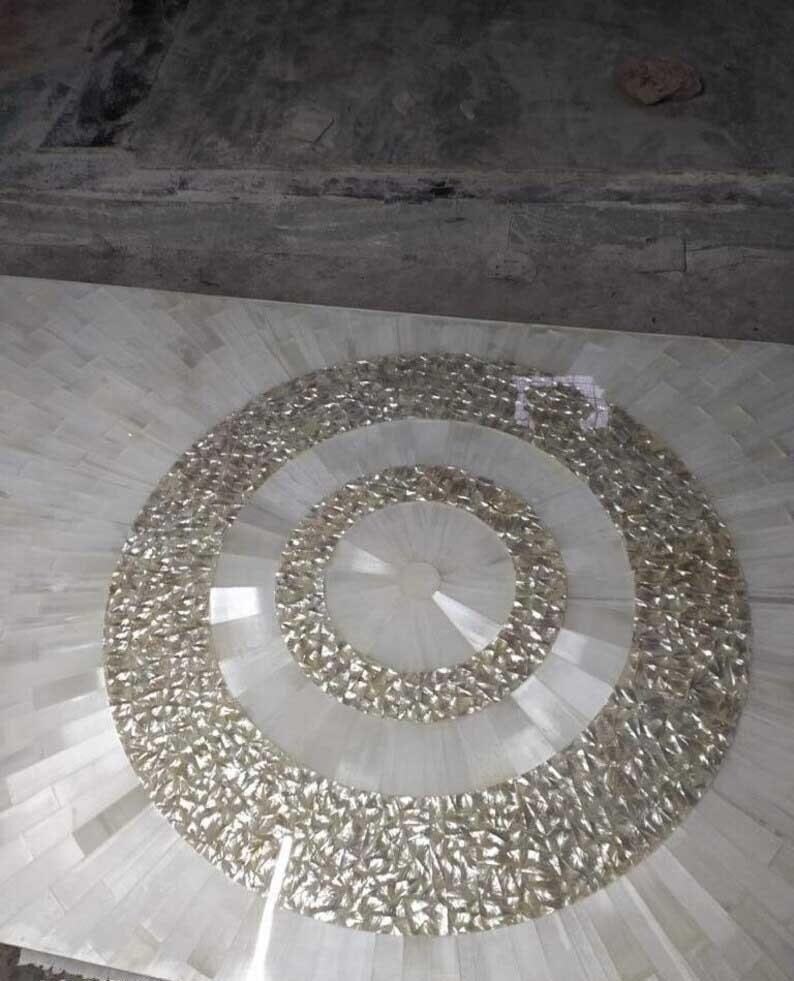 Puede incluir: Una mesa de mosaico con un dise&ntilde;o circular. El centro presenta anillos conc&eacute;ntricos de material brillante e iridiscente. La parte exterior de la mesa est&aacute; hecha de baldosas rectangulares blancas, que irradian hacia afuera desde el centro.