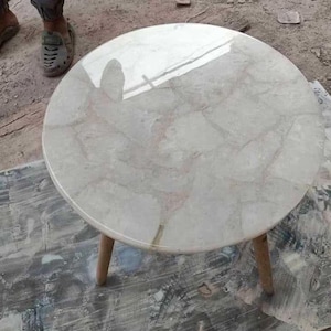 May include: A round side table with a light-colored, patterned top and three wooden legs. The tabletop has a glossy finish with a marbled appearance, featuring shades of white, gray, and hints of pink. The legs are a light brown color.