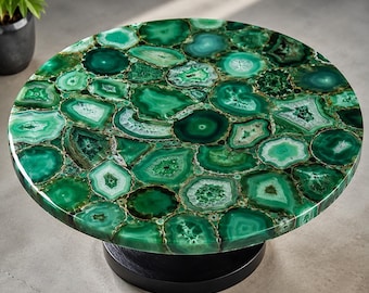 Può includere: Un tavolino rotondo con un piano composto da numerose fette di agata verde lucidata disposte a mosaico. La superficie presenta varie tonalità di verde smeraldo e bosco con venature naturali, sostenuto da una base cilindrica nera.