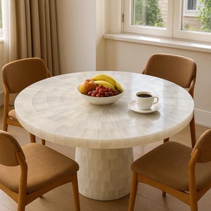 Puede incluir: Una mesa de comedor redonda con una tapa de mosaico de azulejos blancos y una base de pedestal. La mesa está rodeada por cuatro sillas tapizadas marrones. Un cuenco de fruta y una taza de café están sobre la mesa. La mesa tiene unos 122 cm de diámetro.