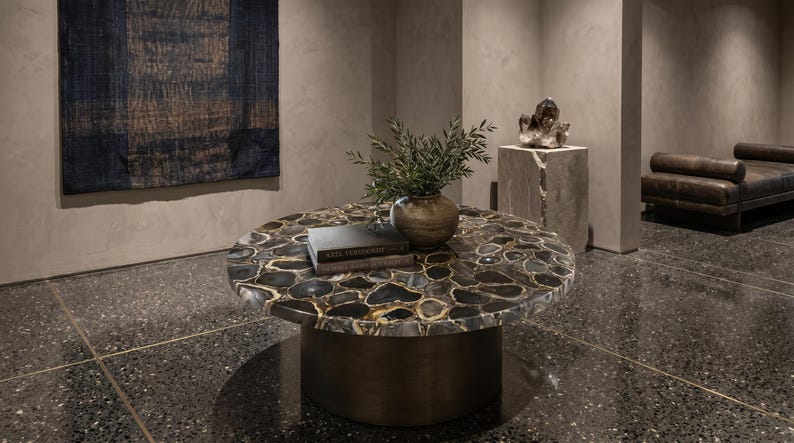 May include: A round coffee table featuring a top made of dark, polished agate slices with gold-toned veining, supported by a solid cylindrical base. A ceramic vase with green foliage and two stacked books rest on the table surface.