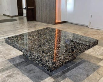 May include: A square coffee table with a dark, patterned surface. The table has a low, rectangular base and a polished, reflective top. The surface features a mosaic-like design with black and cream-colored elements. The table is a statement piece for a living room.