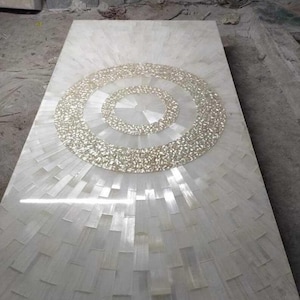 Puede incluir: Una mesa rectangular de piedra blanca con un dise&ntilde;o circular texturizado en el centro. El dise&ntilde;o est&aacute; hecho de peque&ntilde;as piezas de color claro, y la superficie de la mesa tiene un patr&oacute;n radiante.