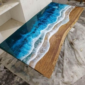 Puede incluir: Una mesa rectangular de madera con una superficie de resina, dise&ntilde;ada para parecerse a una escena de playa. La parte superior presenta un degradado de azules para el oc&eacute;ano, blanco para las olas y una veta de madera natural para la arena. La mesa mide unos 152 cm de largo.
