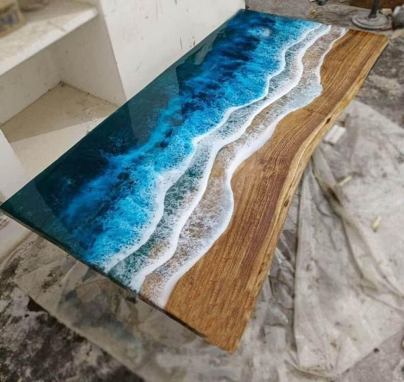 Puede incluir: Una mesa rectangular de madera con una superficie de resina, dise&ntilde;ada para parecerse a una escena de playa. La parte superior presenta un degradado de azules para el oc&eacute;ano, blanco para las olas y una veta de madera natural para la arena. La mesa mide unos 152 cm de largo.