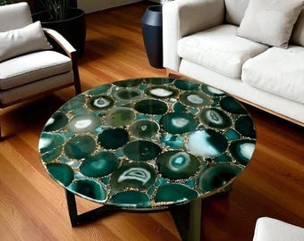 May include: Round coffee table with a top made of green agate stones with gold accents. The table has a dark base and is set in a living room with a sofa and chairs.