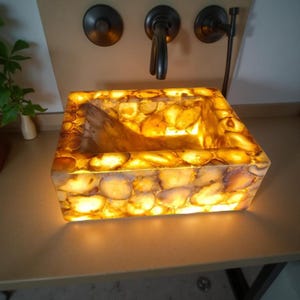May include: A rectangular, backlit sink made of amber-colored, polished stones. The sink has a built-in basin and is set against a neutral-toned wall with dark bronze fixtures. The sink's interior is a lighter shade of amber.