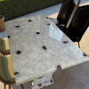 Puede incluir: Una mesa rectangular de piedra blanca con detalles en negro. La mesa tiene una superficie texturizada única y está sostenida por una base de pedestal. Dos sillas de cuero negro están colocadas detrás de la mesa, con un seto verde al fondo.