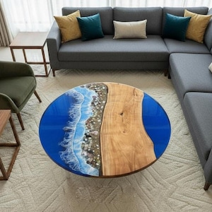 Puede incluir: Una mesa de centro redonda con un diseño único. La mesa presenta una tapa de resina epoxi azul con un diseño de olas y playa en un lado y madera natural en el otro. La mesa está rodeada por un sofá seccional gris.