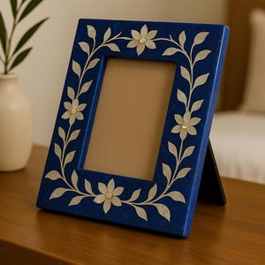 Könnte beinhalten: Ein rechteckiger blauer Bilderrahmen mit floralem Design. Der Rahmen hat eine dunkelblaue Oberfläche mit einem weißen Blumen- und Blättermuster. Der Rahmen steht auf einer Holzoberfläche.