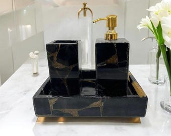 Può includere: Un set di accessori da bagno neri e dorati. Il set include un dosatore di sapone con pompa dorata, un bicchiere e un vassoio. Gli articoli hanno un aspetto marmorizzato con venature dorate. Sono visibili anche una bottiglia di vetro trasparente e un vaso di fiori bianchi.