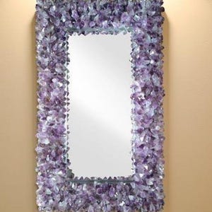 Op de afbeelding: Rechthoekige spiegel met een frame volledig bedekt met amethistkristallen. De kristallen, in verschillende tinten paars en lavendel, vormen een getextureerde en sprankelende rand rond het reflecterende glasoppervlak.