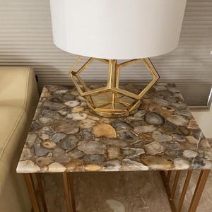May include: A side table with a mosaic stone top and gold metal legs. A gold geometric lamp base supports a white lampshade. The table's surface is a mix of neutral colors, including gray, brown, and beige.