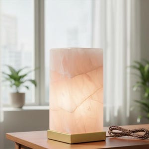 K&ouml;nnte beinhalten: Rechteckige, beleuchtete Lampe aus rosa Onyx mit goldenem Sockel. Die Lampe steht auf einem Holztisch, mit einem braunen Kabel. Im Hintergrund sind ein Fenster, Vorh&auml;nge und eine Topfpflanze zu sehen.