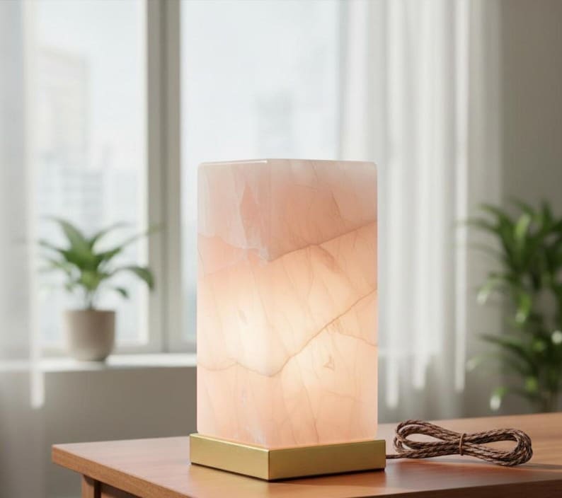 K&ouml;nnte beinhalten: Rechteckige, beleuchtete Lampe aus rosa Onyx mit goldenem Sockel. Die Lampe steht auf einem Holztisch, mit einem braunen Kabel. Im Hintergrund sind ein Fenster, Vorh&auml;nge und eine Topfpflanze zu sehen.