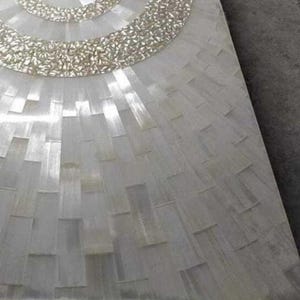 Puede incluir: Primer plano de un dise&ntilde;o de mosaico rectangular blanco. El mosaico presenta un patr&oacute;n circular en la parte superior con un borde texturizado de color plateado. El resto del azulejo est&aacute; formado por piezas rectangulares.