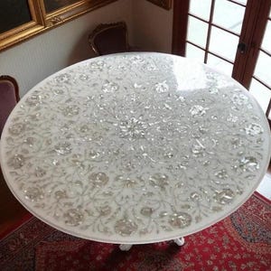 May include: Oval white marble table with floral inlay design. The table has a polished surface and rests on three white legs. The table is approximately 60 inches in diameter.