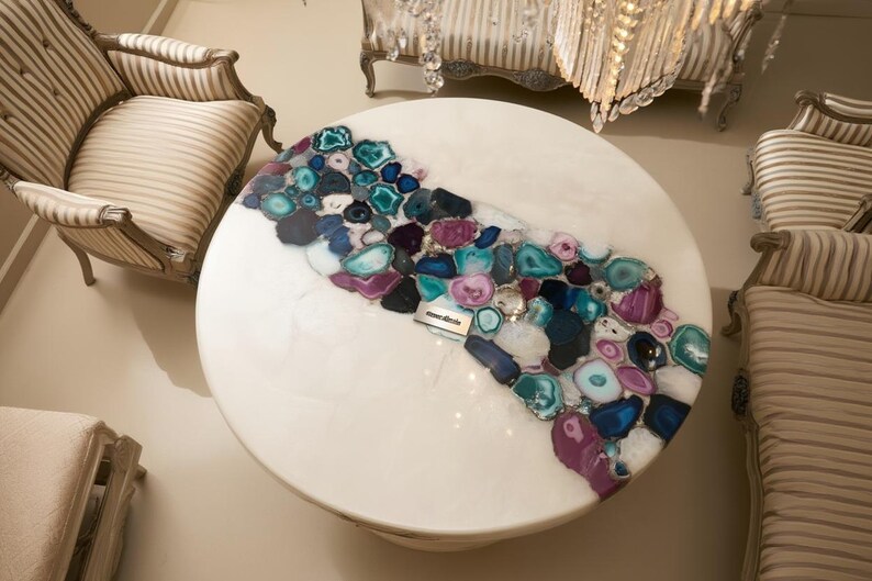 May include: A round white coffee table with a decorative inlay of colorful agate stones in shades of blue, purple, and teal. The table is surrounded by striped upholstered furniture, creating a luxurious living room setting.