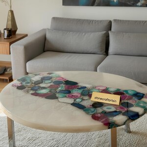 May include: Round coffee table with a cream-colored surface and a river of colorful agate stones. The table has silver metal legs and a gold tag that reads "stonecraftartsin".