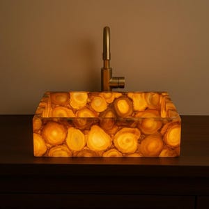 May include: A rectangular, illuminated sink basin with a unique, patterned surface. The sink is made of a translucent, amber-coloured material, with a gold-toned tap. The sink is set on a dark brown surface.