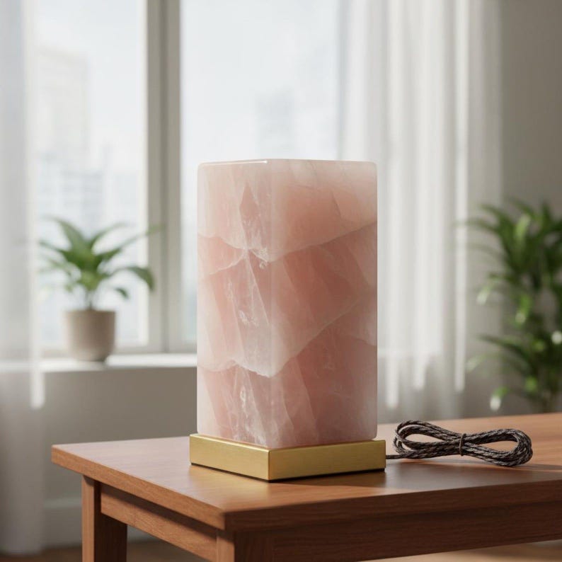 K&ouml;nnte beinhalten: Eine rechteckige, zartrosa, durchscheinende Steinlampe mit goldfarbenem Sockel steht auf einem Holztisch. Die Lampe hat eine zylindrische Form und eine strukturierte Oberfl&auml;che. Ein geflochtenes Netzkabel ist sichtbar. Eine Topfpflanze ist im Hintergrund.