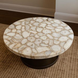 Puede incluir: Una mesa de centro redonda con una tapa de mosaico de piedra blanca y beige. La mesa tiene una base cilíndrica marrón oscuro y está sobre una alfombra de color neutro. La tapa de piedra tiene un patrón natural único.
