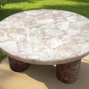 May include: A round coffee table with a light pink and white mosaic top. The table is supported by two cylindrical legs. The table is outdoors on a sunny day.