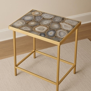 May include: A rectangular side table with a gold-coloured metal frame. The tabletop is made of polished agate stones in shades of grey, white, and brown. The table has a simple, geometric design and is suitable for a living room or bedroom.