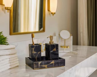 Può includere: Un set di accessori per il bagno neri e dorati, tra cui un portaspazzolino, un dosatore di sapone e un vassoio. Il set è realizzato in un materiale scuro con venature dorate. Uno spazzolino dorato è nel supporto.