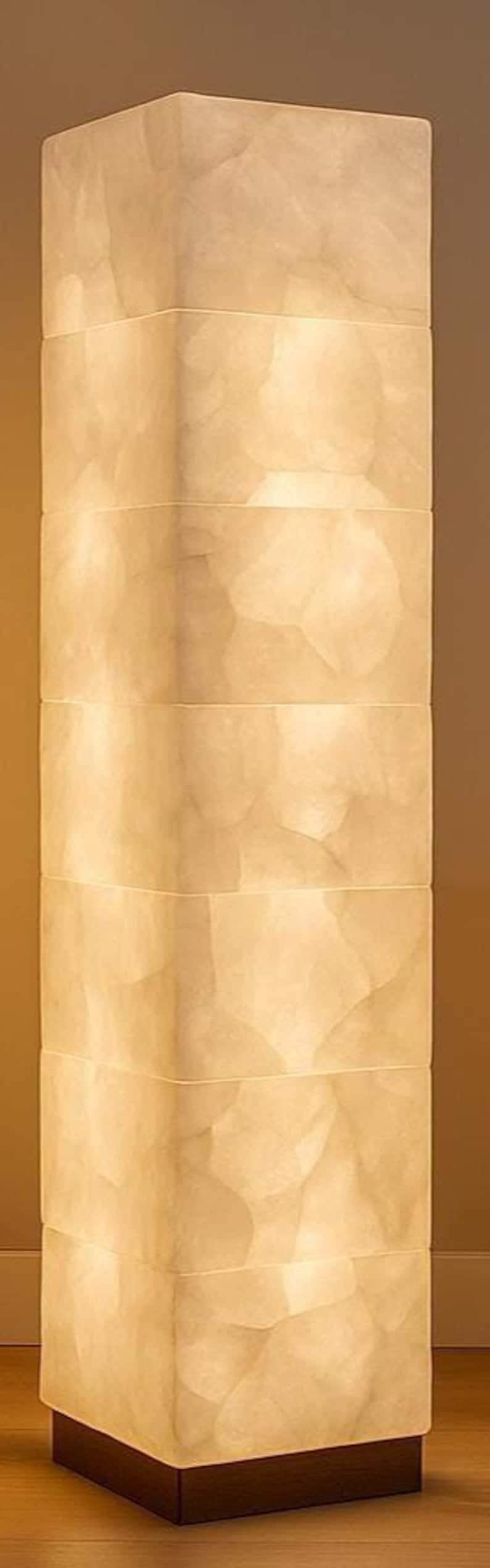 May include: A tall, rectangular floor lamp with a warm, glowing light. The lamp is made of a light-colored, textured material, possibly alabaster or a similar stone. The base is a dark brown color.
