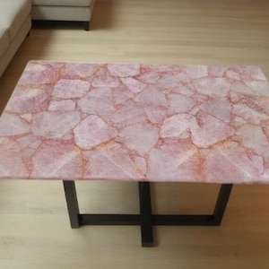 May include: Rectangular coffee table with a rose quartz top. The tabletop is made of irregular, light pink stone pieces. The table has a dark, geometric metal base. The table is approximately 122 cm long and 61 cm wide.
