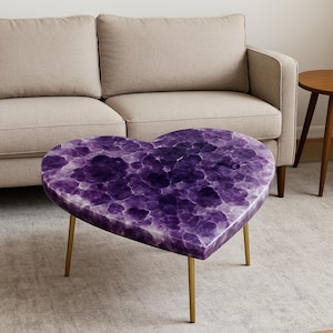 May include: A heart-shaped coffee table with a purple amethyst design top and gold-colored legs. The table is placed in front of a beige sofa and a small wooden side table, creating a cozy living room setting.